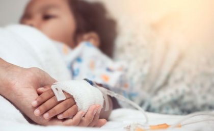 An adult hand holds a bandaged child's hand in the foreground, the child lays in a hospital bed.